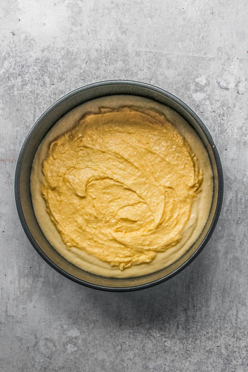 Almond frangipane sits on top of an almond cake batter in a round metal tin on a gray surface.