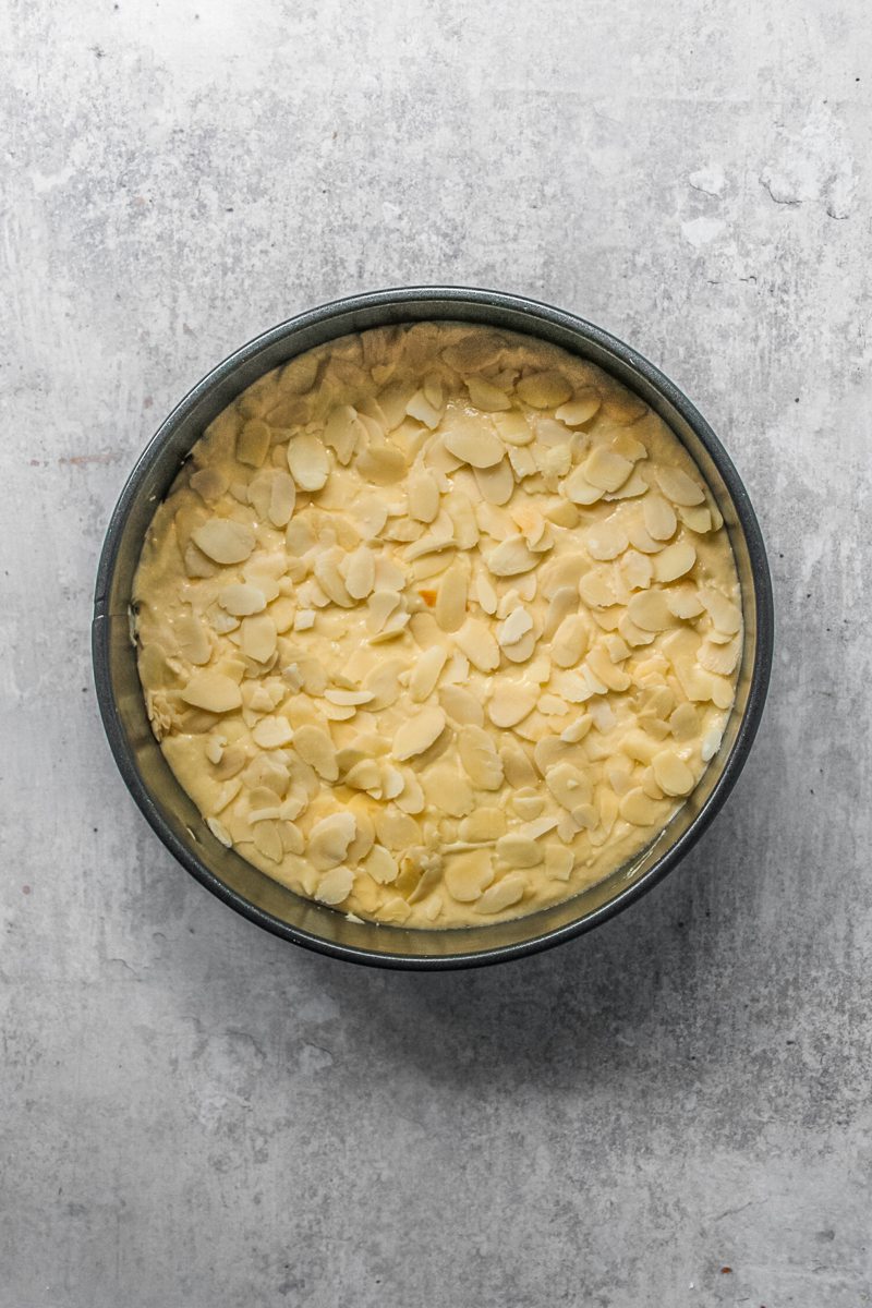 Sliced almonds sit on top of unbake almond cake batter in a round metal tin on a gray surface.