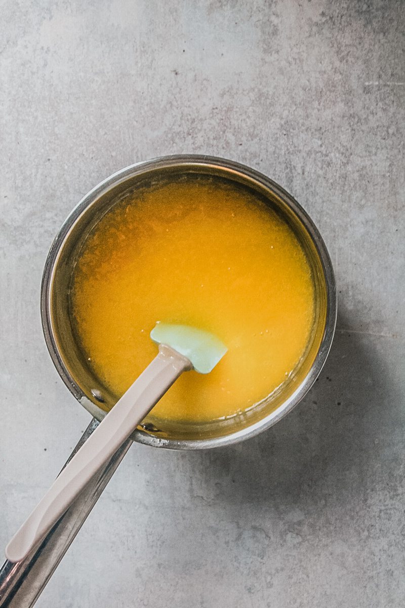 A butter and cream mixture sits in a stainless steel saucepan with a spatula resting therein on a light gray surface.