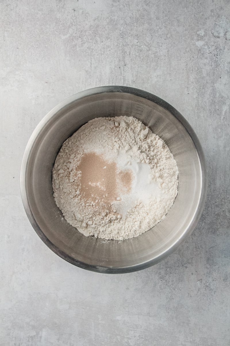 Yeast, sugar and flour sit in a stainless steel bowl on a gray surface.