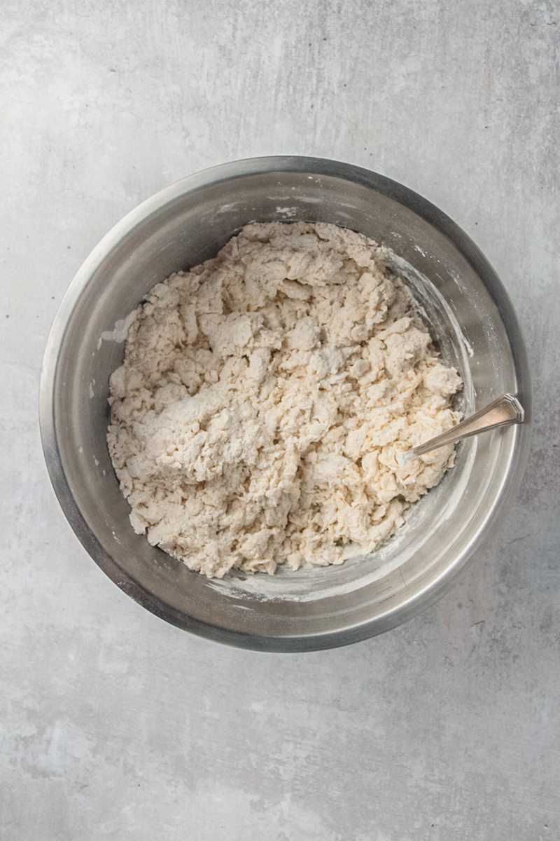 A shaggy dough sits in a stainless steel dough on a gray surface.