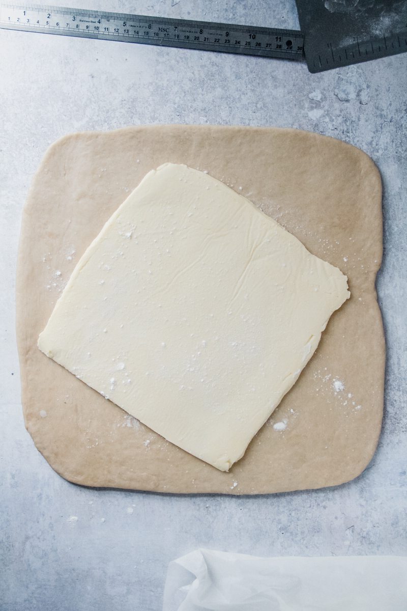 A triangle of butter sits in the center of a square of rolled out dough on a gray surface.