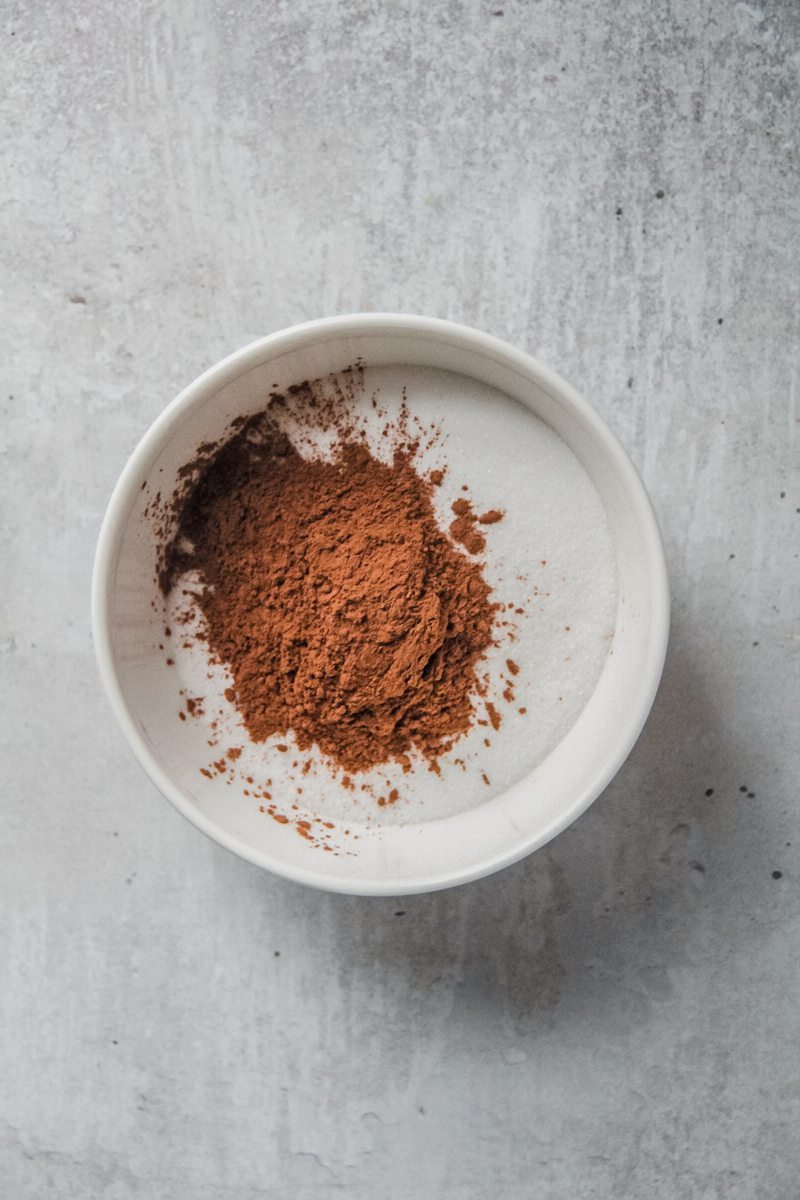 Ground cinnamon sits on top of sugar in a white ceramic bowl on a gray surface.