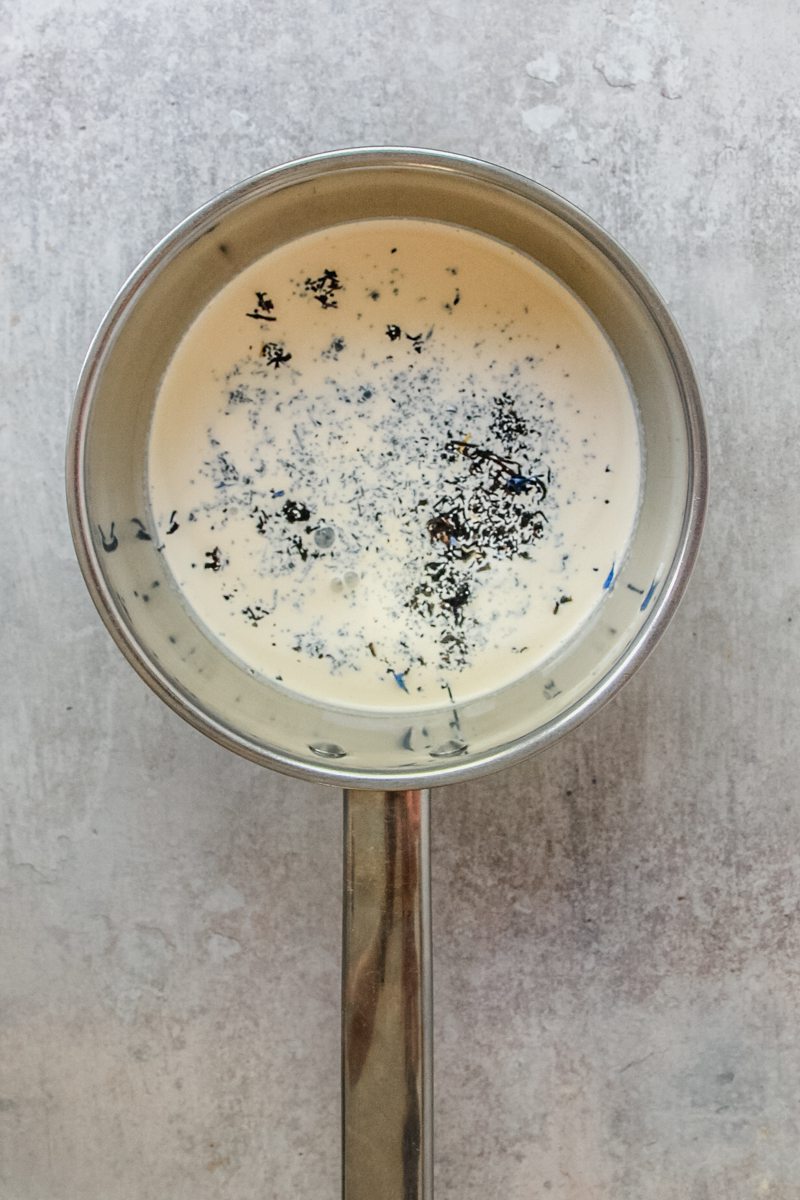 Loose earl gray tea sits on top of heavy cream in a small stainless steel saucepan on a gray surface.