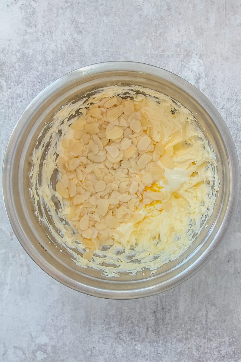 Whipped butter and sugar sits in a stainless steel bowl with flaked almonds and finely chopped lemon zest on top on a gray surface.