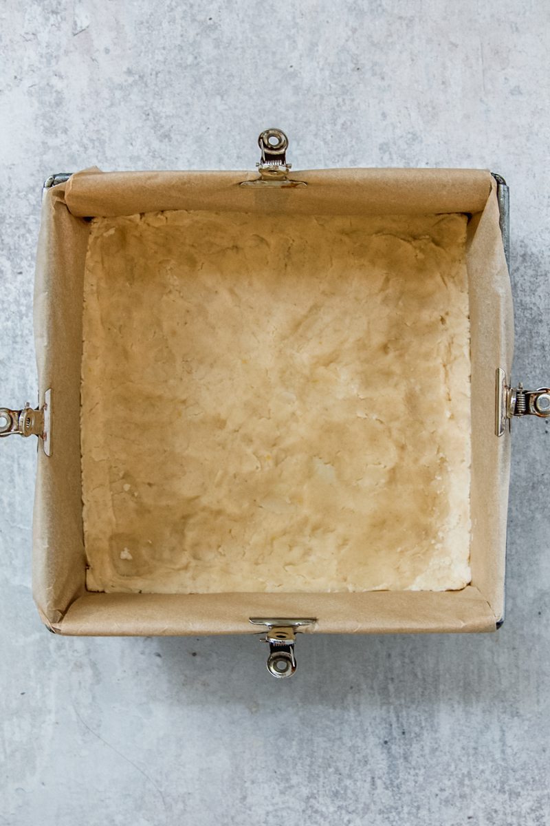 A shortbread style crust sits pressed into a lined square metal baking tin on a gray surface.