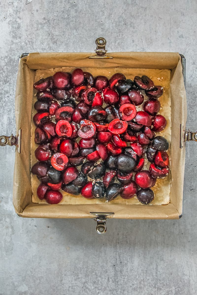 Halved cherries sit on top of a shortbread style base pressed into a lined square metal baking tin on a gray surface.