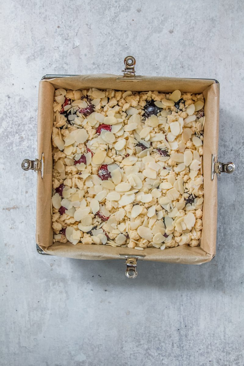 A crumb topping and flaked almonds sit on top of halved pitted cherries in a lined square metal baking tin on a gray surface.