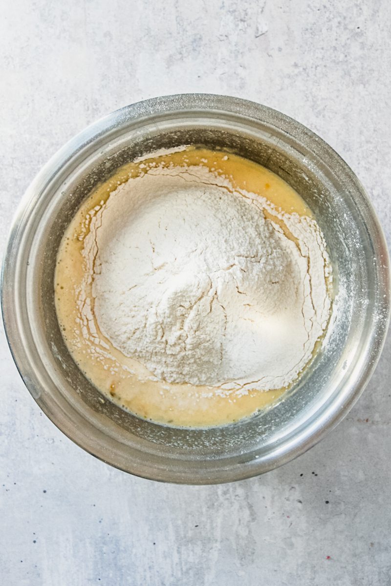 Dry ingredients sit on top of a pancake mixture in a stainless steel bowl on a gray surface.