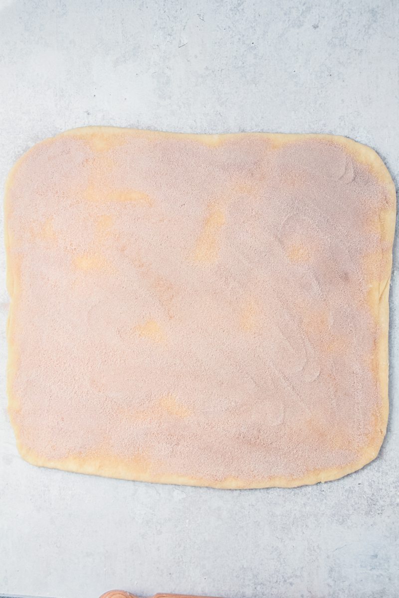 A square of dough with a cinnamon sugar mixture on top sits on a gray surface.