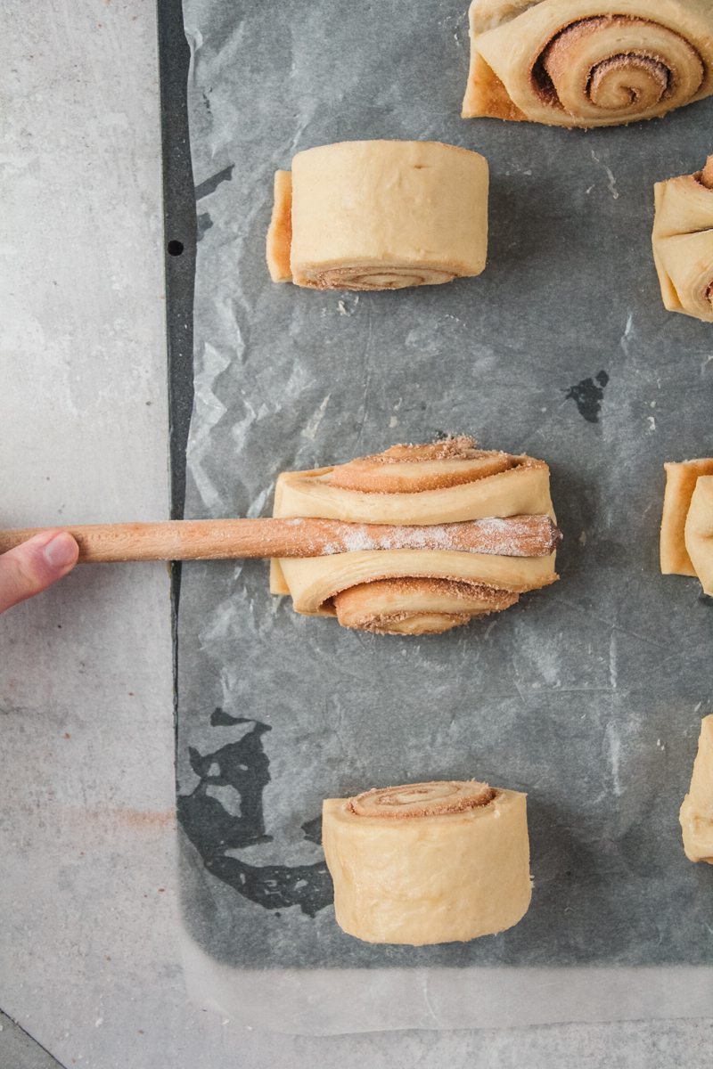 A cinnamon roll is pressed in the center with the flour coated end of a wooden spoon to create an indent.