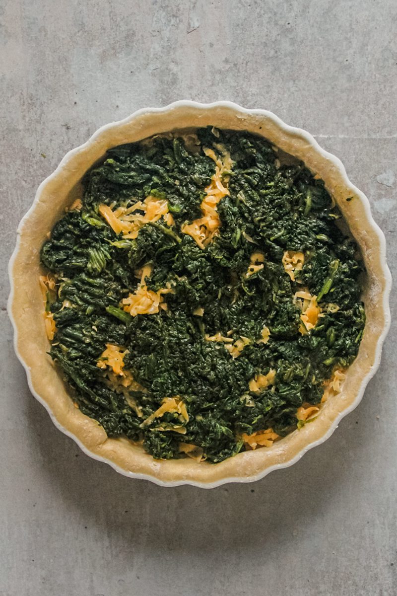 Grated cheese and spinach sit in a quiche pastry in a tart dish on a gray surface.