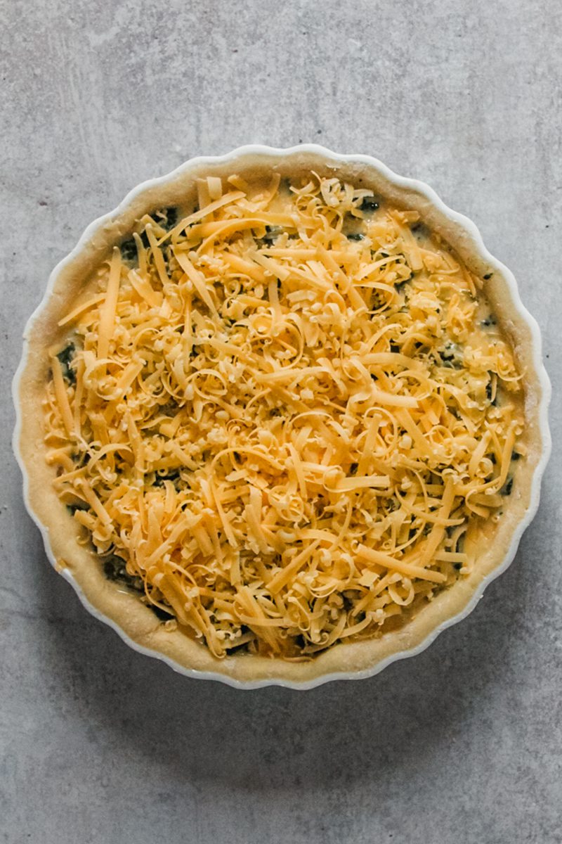 Unbaked spinach and cheese quiche sit in a ceramic tart baking dish on a gray surface.