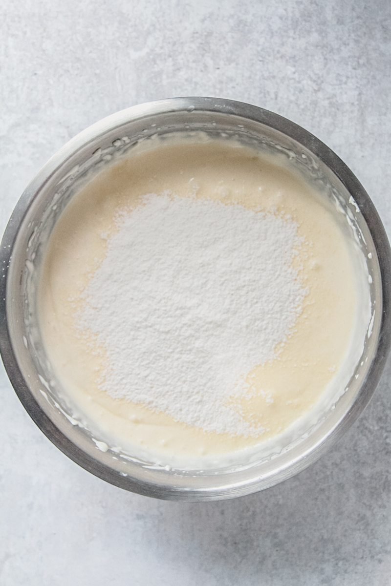 Sifted flour sits on top of a cheesecake mixture in a stainless steel bowl on a gray surface.