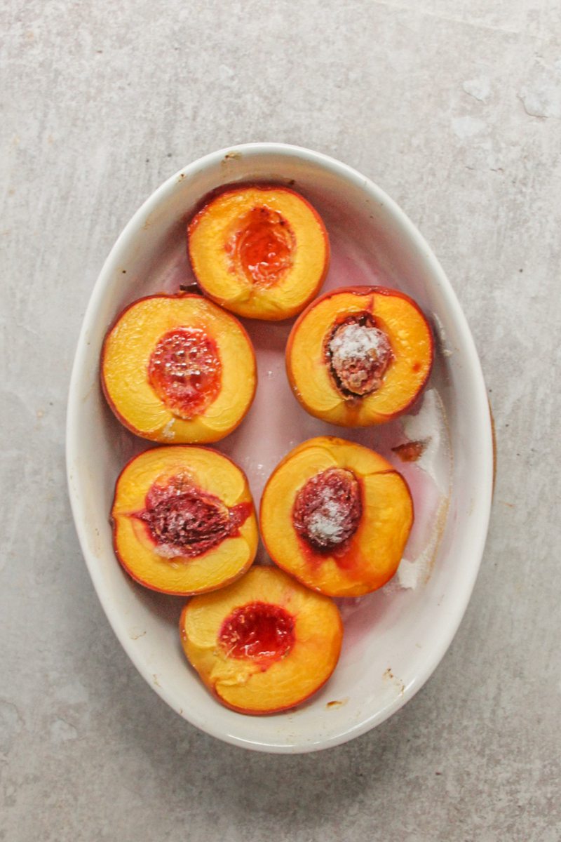 Roasted peaches sit in an oval white ceramic baking dish on a gray surface.