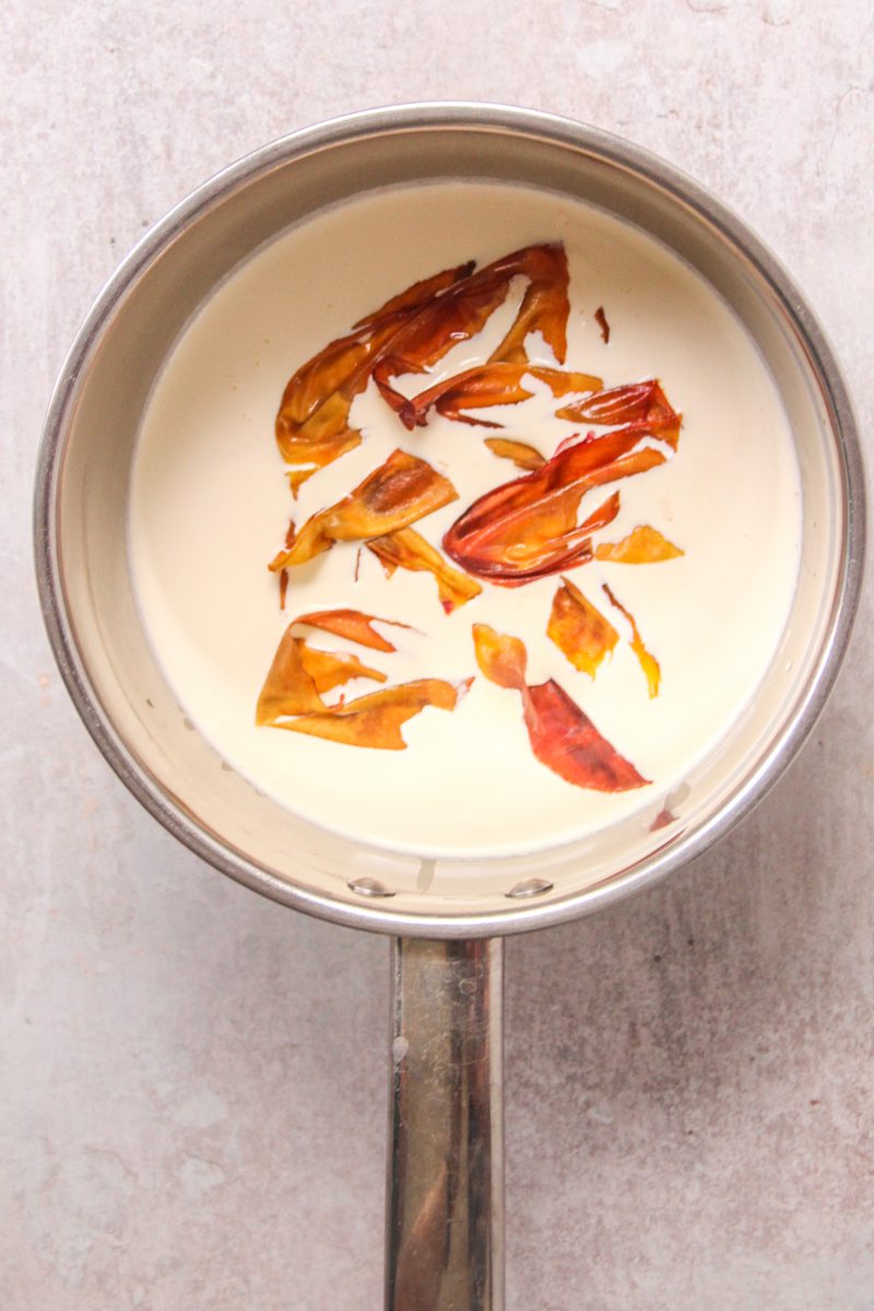Roasted peach skins sit in heavy cream in a stainless steel saucepan on a gray surface.