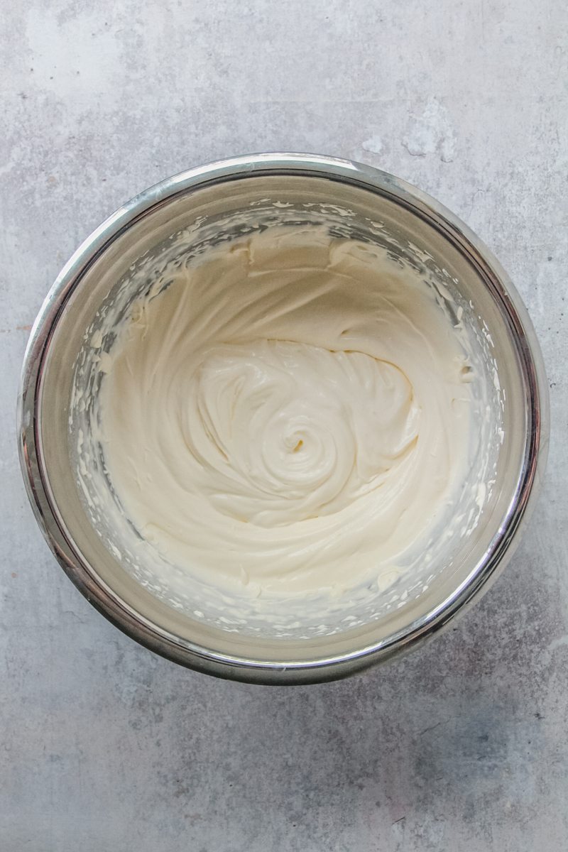 Soft peaks whipped cream sits in a stainless steel bowl on a gray surface.