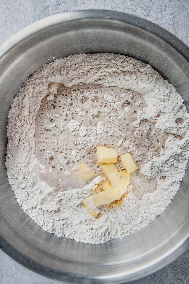 Flour and butter sit on top of bloomed yeast in a stainless steel bowl on a gray surface.