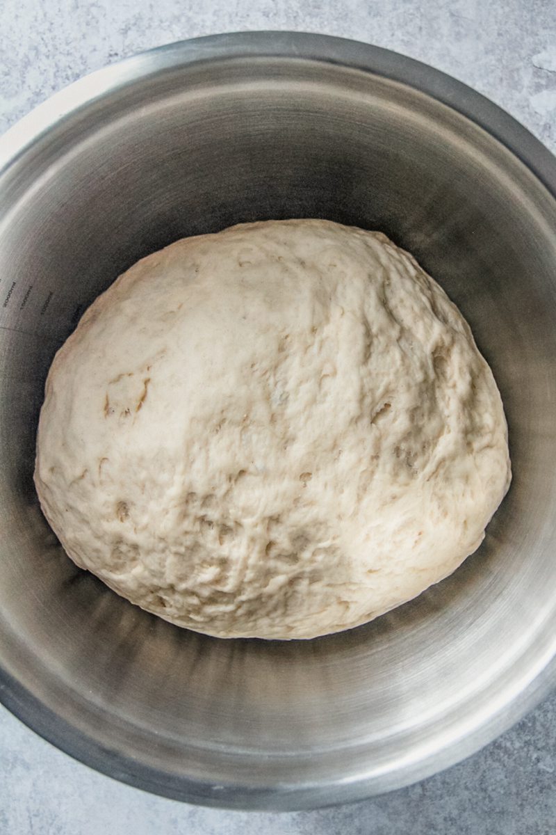 A ball of smooth yeast dough sits in a stainless steel bowl on a gray surface.