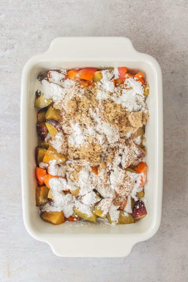 Cubes of plums and apricots sit in a rectangle white ceramic baking dish with a sugar and cornflour mixture on top on a gray surface.