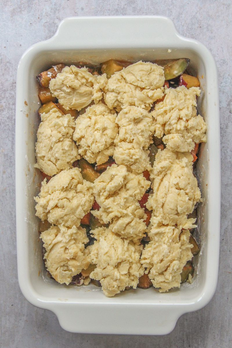Almond biscuits sit on top of a plum and apricot mixture in a white ceramic rectangle baking dish on a gray surface.