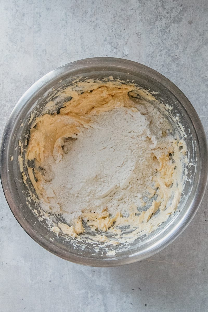 Flour sits on top of whipped butter and egg cookie dough in a stainless steel bowl on a gray surface.