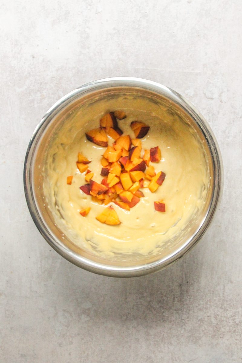 Cubed peach sits on top of cake batter in a stainless steel bowl on a gray surface.