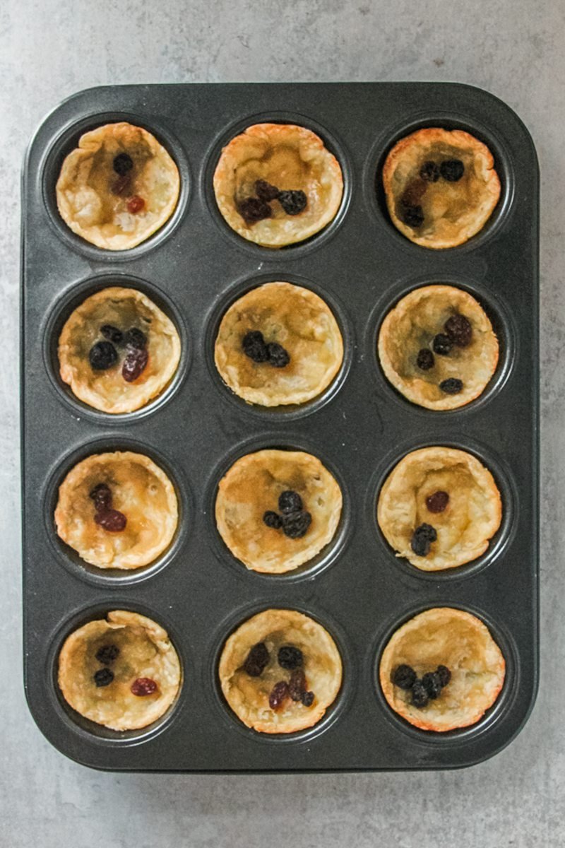 Blind baked butter tart pastry cases with a few raisins in each sit in a muffin tin on a gray surface.