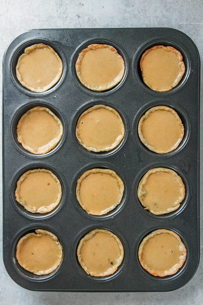 Butter tart pastry cases are filled with the unbaked syrup center in a muffin tin on a gray surface.