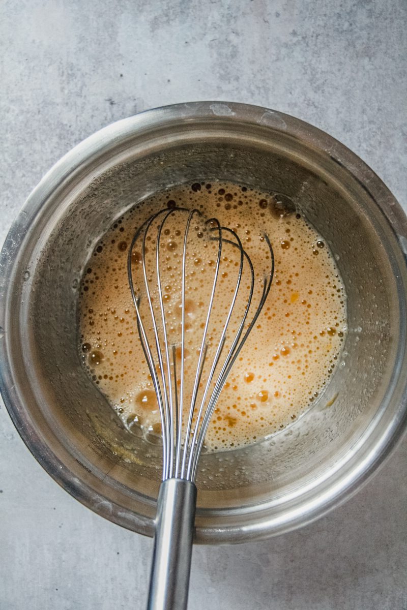 Whisked eggs sit in a stainless steel bowl with a whisk resting therein on a gray surface.