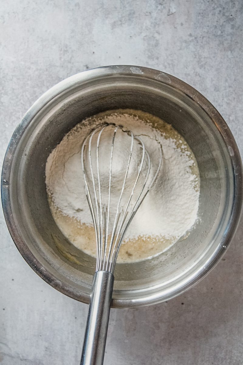 Flour sits on top of whisked eggs and sugar in a stainless steel bowl with a whisk resting therein on a gray surface.