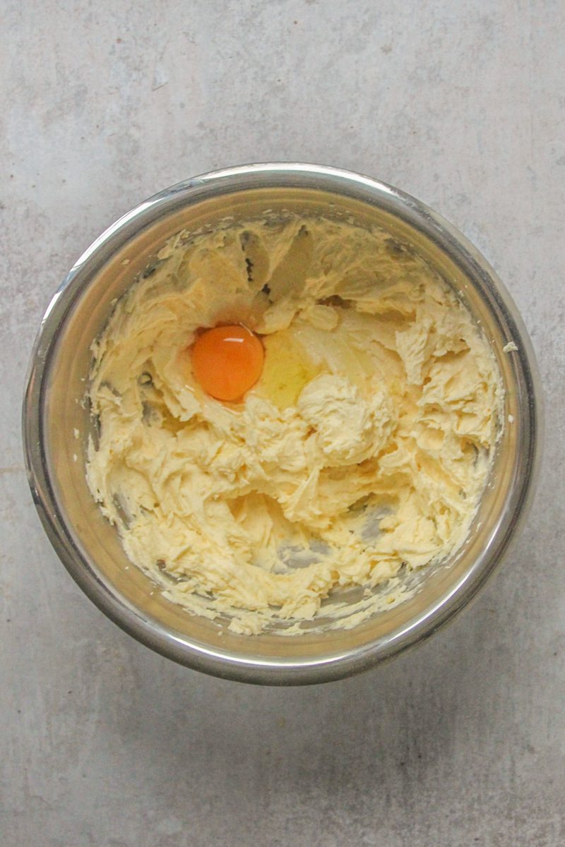 An egg sits on top of whipped butter and sugar in a stainless steel bowl on a gray surface.