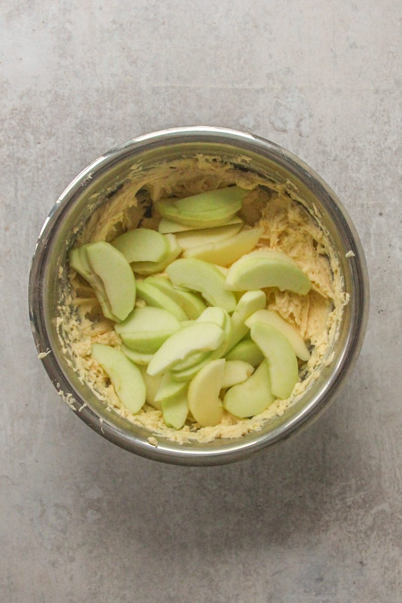 Apple splices sit on top of cake batter in a stainless steel bowl on a gray surface.