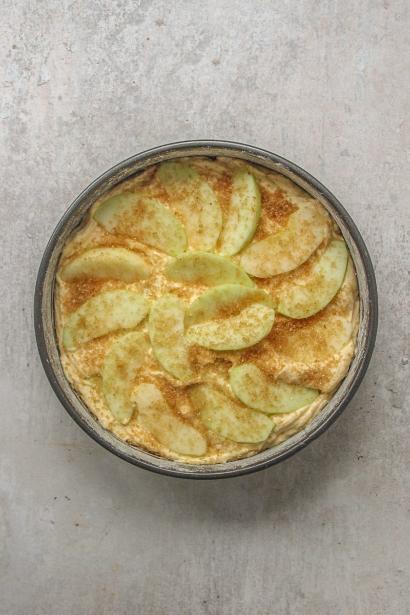Apple slices and demerara sugar sit on top of cake batter in round metal cake tin on a gray surface.