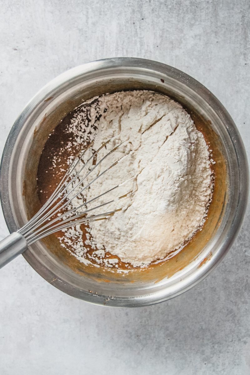 Dry ingredients sit on top of sweet potato cake batter in a stainless steel bowl on a gray surface.