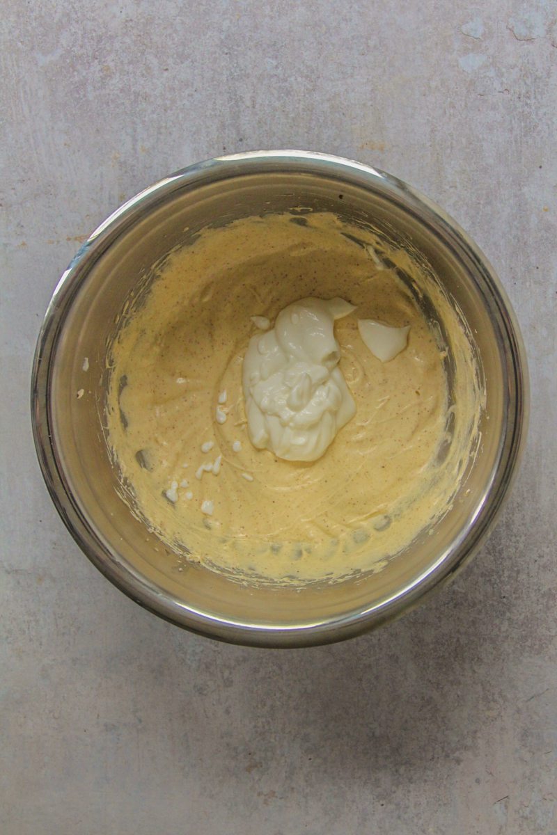 Yogurt sits on top of a whipped butter, sugar and egg micture in a stainless steel bowl on a gray surface.