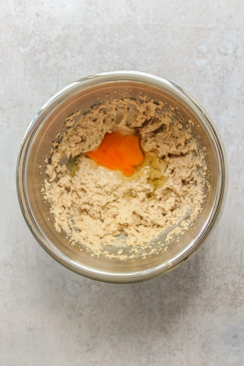 An egg sits on top of whipped brown butter and sugar in a stainless steel bowl on a gray surface.