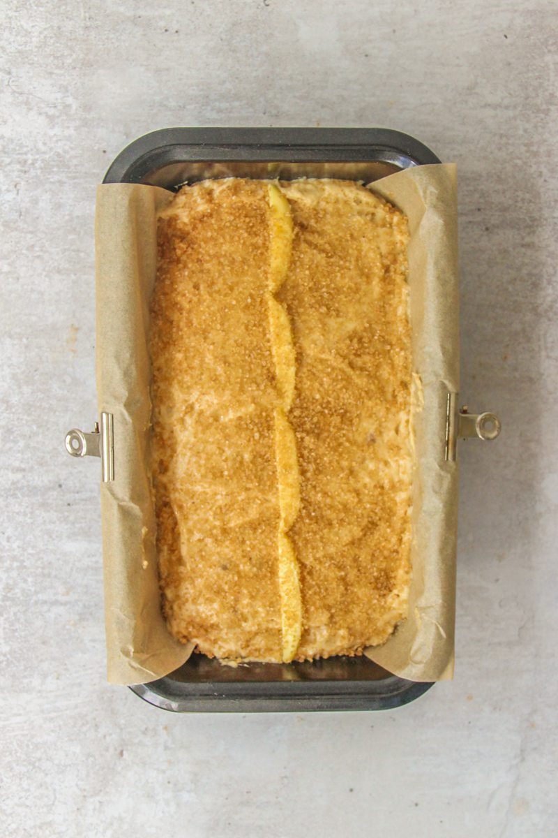 Apple butter batter sits in a loaf tin with demerara sugar and a line of butter on top in a black metal tin on a gray surface.