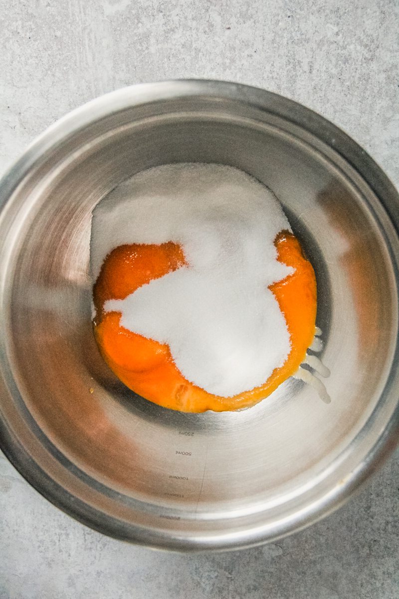 Sugar and egg yolks sit in a stainless steel bowl on a gray surface.