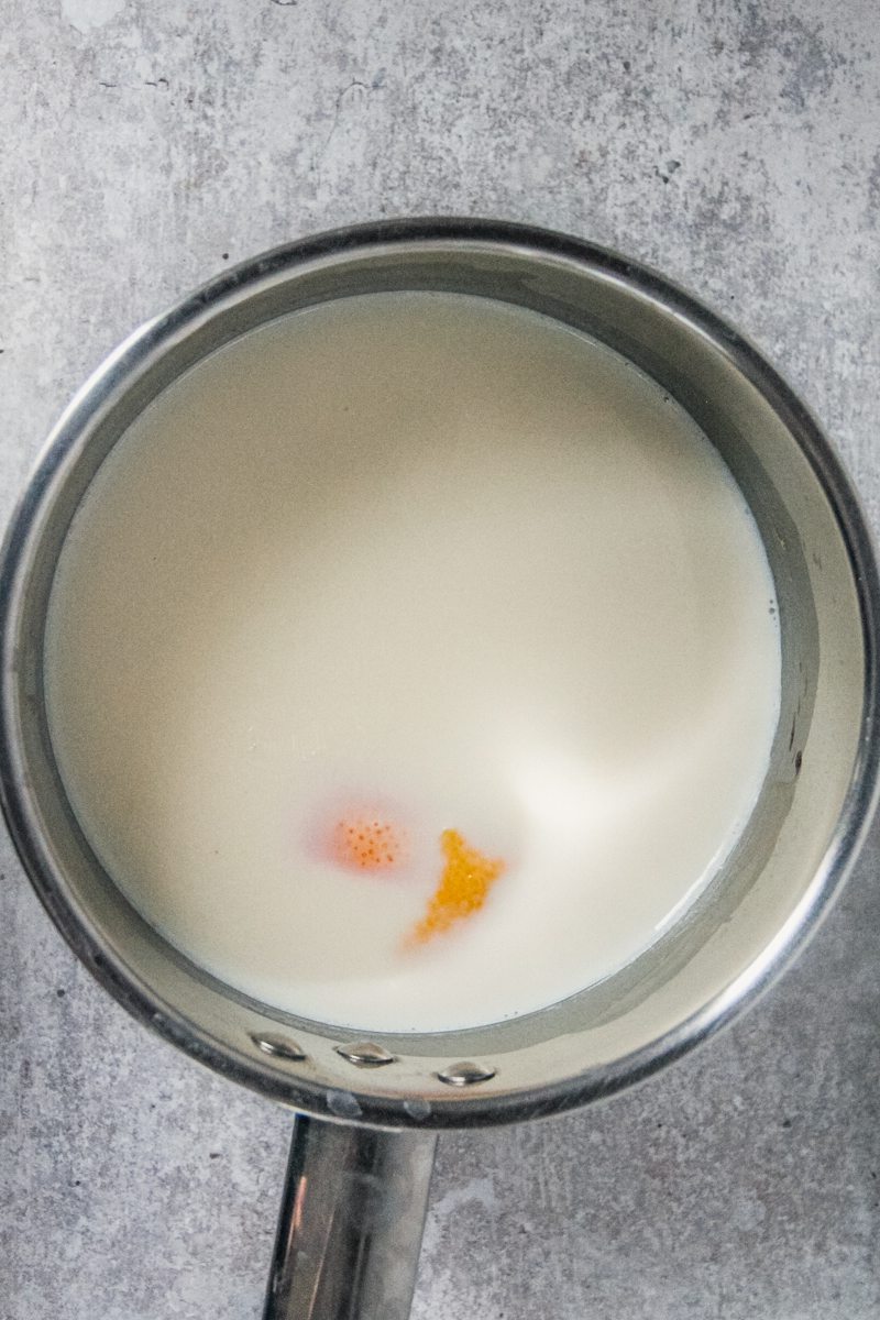 Milk and citrus peels sit in a stainless steel saucepan on a gray surface.