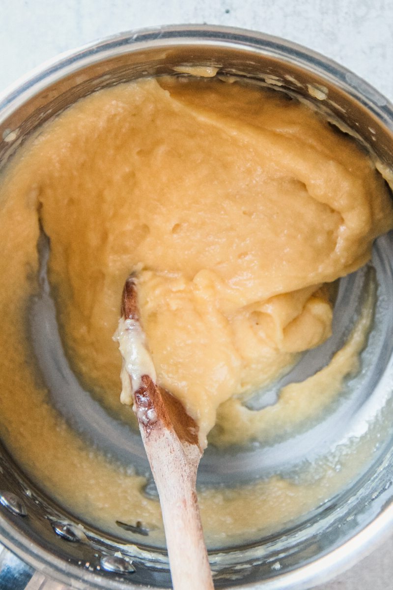 Choux pastry sits in a stainless steel saucepan with a wooden spoon on a gray surface.