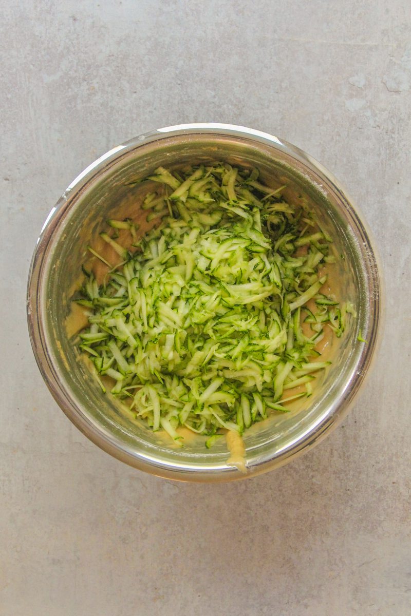 Grated zucchini sits on top of a cake batter in a stainless steel bowl on a gray surface.