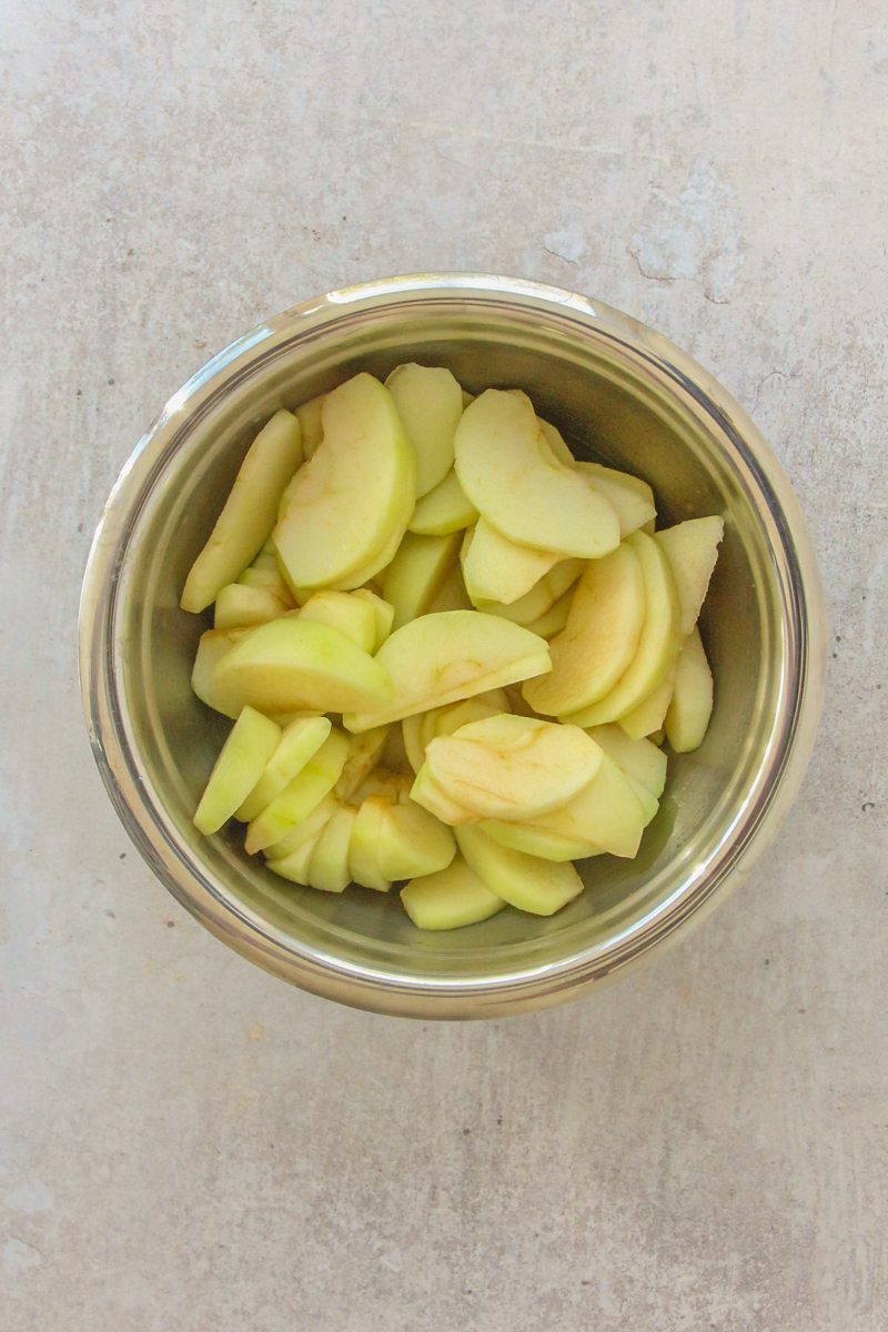 Slices of apples sit coated in lemon juice in a stainless steel bowl on a gray surface.
