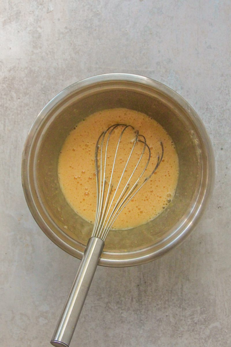Whisked eggs, sugar and flour sits in a stainless steel bowl on a gray surface.