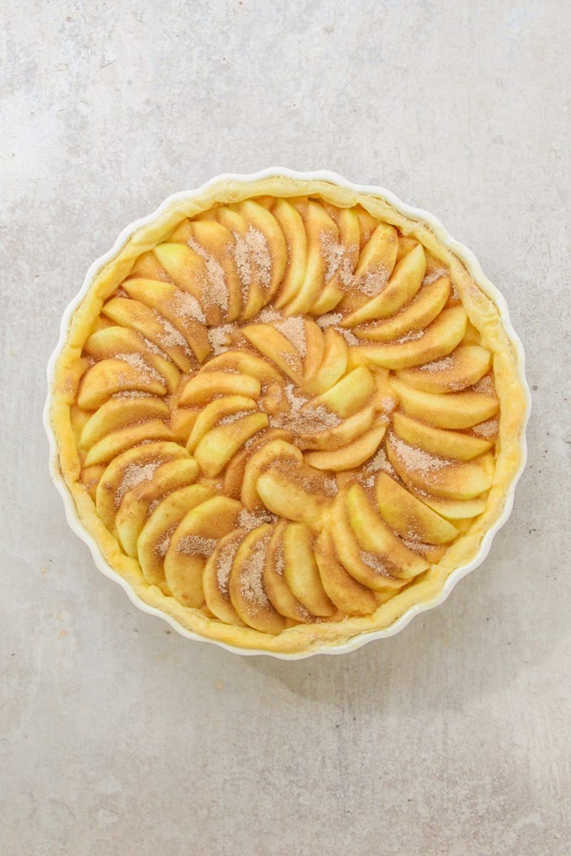 Slices of apple sit laid in a spiral on to top of a custard center and topped with sugar and cinnamon in a pastry case in a white ceramic baking dish on a gray surface.