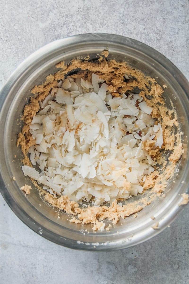 Shaved coconut pieces sit on top of whipped butter and sugar in a stainless steel bowl on a gray surface.