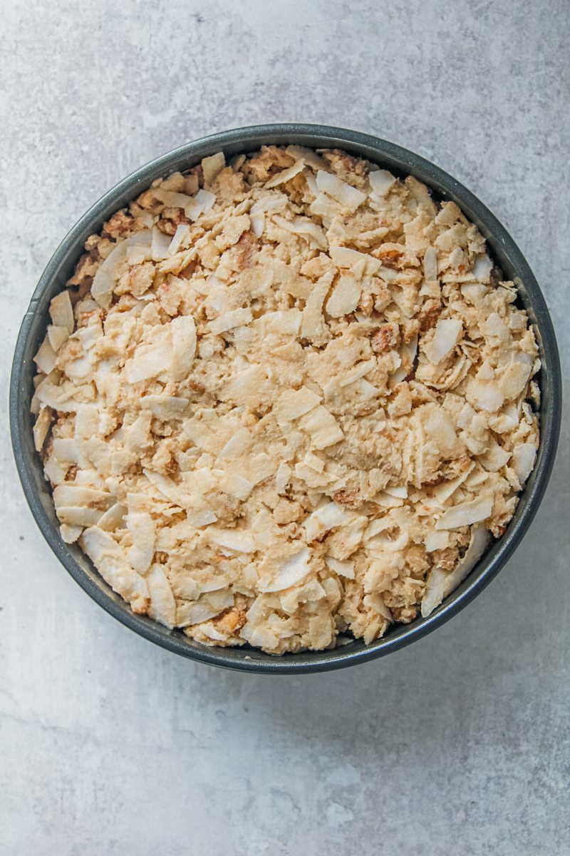 A coconut sugar mixture sits on top of an oatmeal cake in a round cake tin prior to baking on a gray surface.