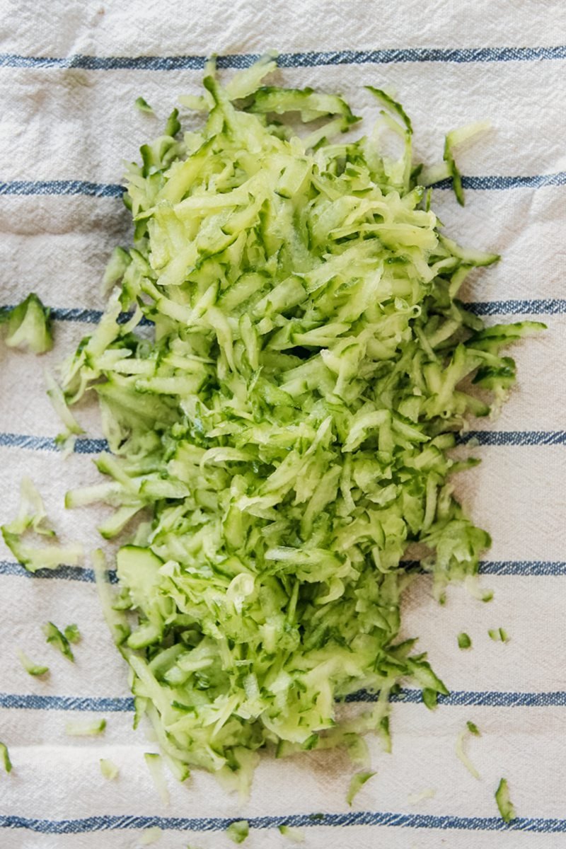 Grated cucumber sits on a clean white and blue striped tea towel to be wrung of water.