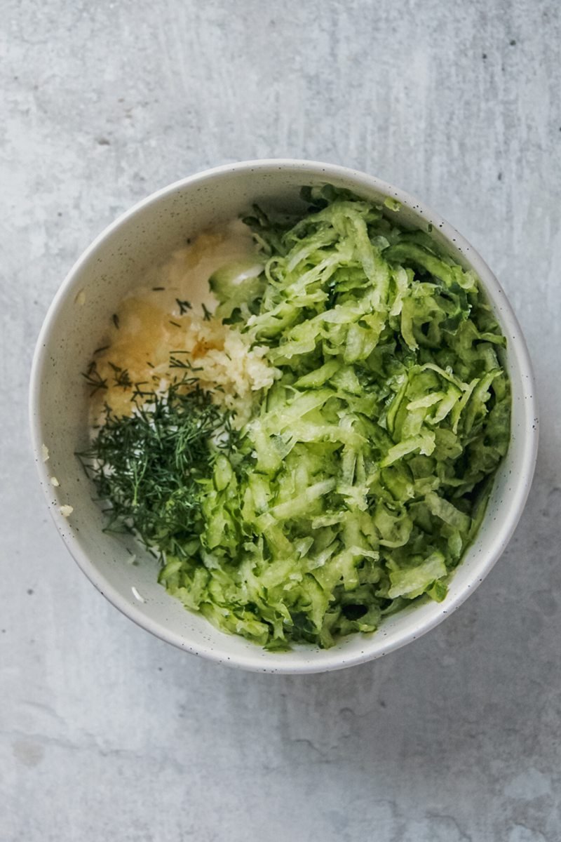 Grated cucumber sits on top of the other ingredients needed for homemade tzatziki on a gray surface.