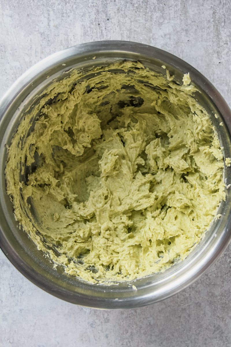 Whipped butter, sugar, egg and pistachio paste sit in a stainless steel bowl on a gray surface.